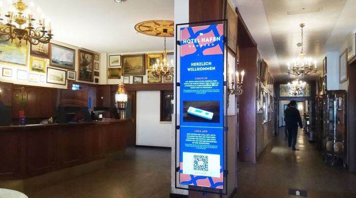 Digital Signage Display im Eingangsbereich des Hotel Hafen mit Begrüßungsinformationen, Check-in-Anleitung, QR-Code zur Nutzung der Luca-App und stilvoller Hotelrezeption im Hintergrund.