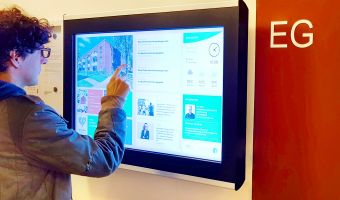 Ein Mann interagiert mit einem digitalen schwarzen Brett, das als Infoboard für Mitarbeiterinformationen dient, mit Inhalten wie Nachrichten, Wetter und internen Updates.