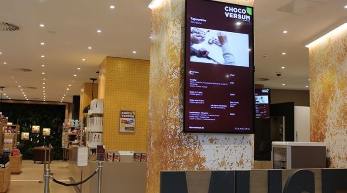 Digital Signage Display im Chocoversum mit Tagesinformationen, Öffnungszeiten und Eintrittspreisen, in einem stilvoll gestalteten Innenraum mit Verkaufsbereich für Schokoladenprodukte.