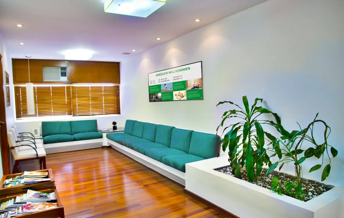 Ein moderner Warteraum mit grünen Sofas und einem digitalen Display an der Wand, das eine Begrüßungsnachricht und Informationen anzeigt.