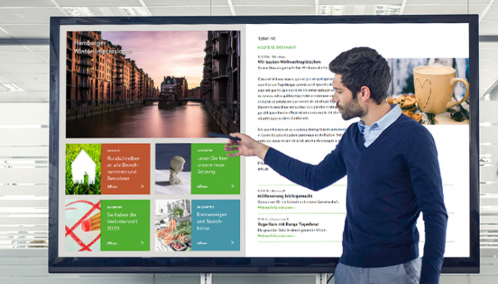 Ein Mann zeigt auf ein digitales schwarzes Brett, das Mitarbeiter mit News, Informationen und Unterhaltungsinhalten versorgt.