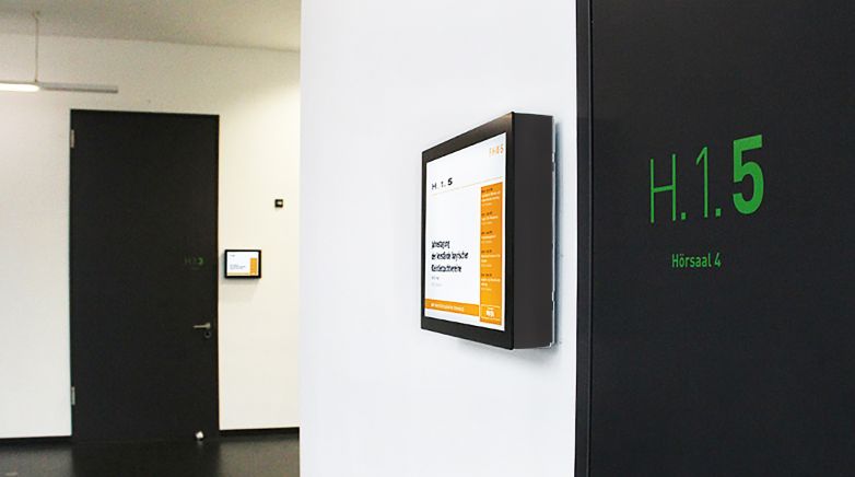 Digitales Türschild zeigt Informationen zu einer Veranstaltung in einem Hörsaal an der Wand eines Konferenzraums.