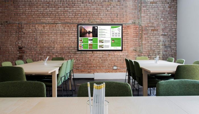 Moderner Besprechungsraum im Büro mit einem digitalen Infoboard an der Wand, das verschiedene Unternehmensinformationen anzeigt. Grüne Stühle und Tische sind für Gruppendiskussionen angeordnet.