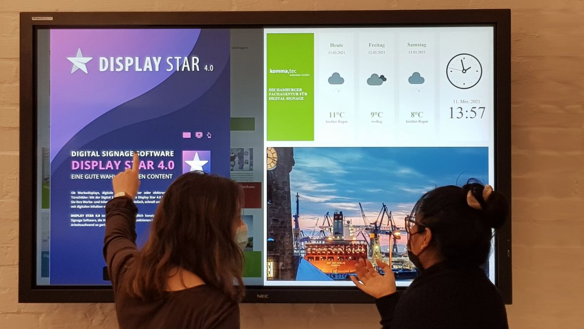 Display Star – Das digitale schwarze Brett für moderne Unternehmenskommunikation Zwei Personen interagieren mit einem digitalen schwarzen Brett, das die Display Star 4.0 Software zeigt, inklusive Wetterinformationen, Uhrzeit und visuellen Inhalten für Unternehmenskommunikation
