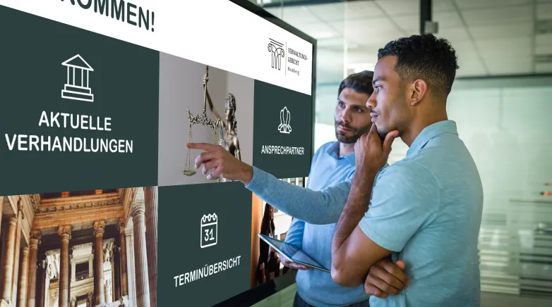 Zwei Männer interagieren mit einem digitalen schwarzen Brett, das Informationen zu aktuellen Verhandlungen, Ansprechpartnern und Terminübersichten anzeigt, in einem modernen Büro.