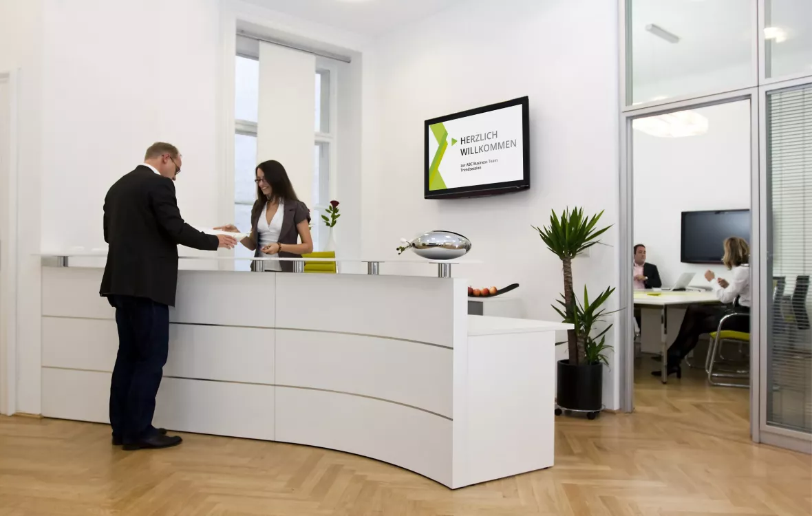 Ein Mann am Empfangsschalter wird von einer Rezeptionistin begrüßt, während ein digitales Display an der Wand eine Begrüßungsnachricht anzeigt.
