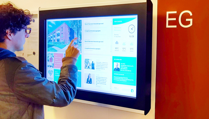 Ein Mann interagiert mit einem digitalen schwarzen Brett, das als Infoboard für Mitarbeiterinformationen dient, mit Inhalten wie Nachrichten, Wetter und internen Updates.