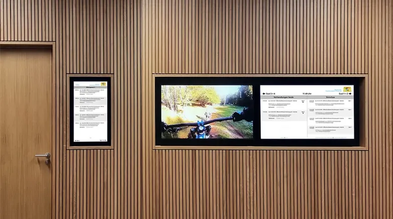 Drei digitale Bildschirme in einer Wand mit Holzvertäfelung, die Informationen, Bilder und Inhalte anzeigen, darunter ein Bild von einem Fahrradfahrer auf einem Waldweg.