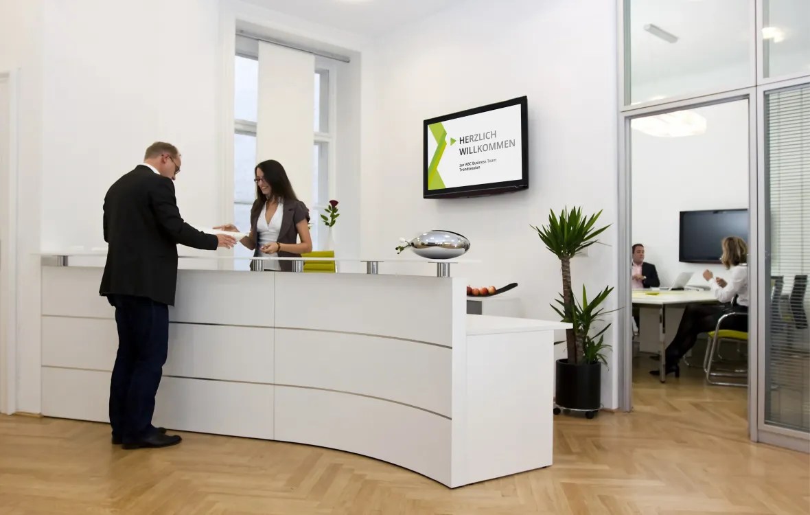 Ein Mann am Empfangsschalter wird von einer Rezeptionistin begrüßt, während ein digitales Display an der Wand eine Begrüßungsnachricht anzeigt.