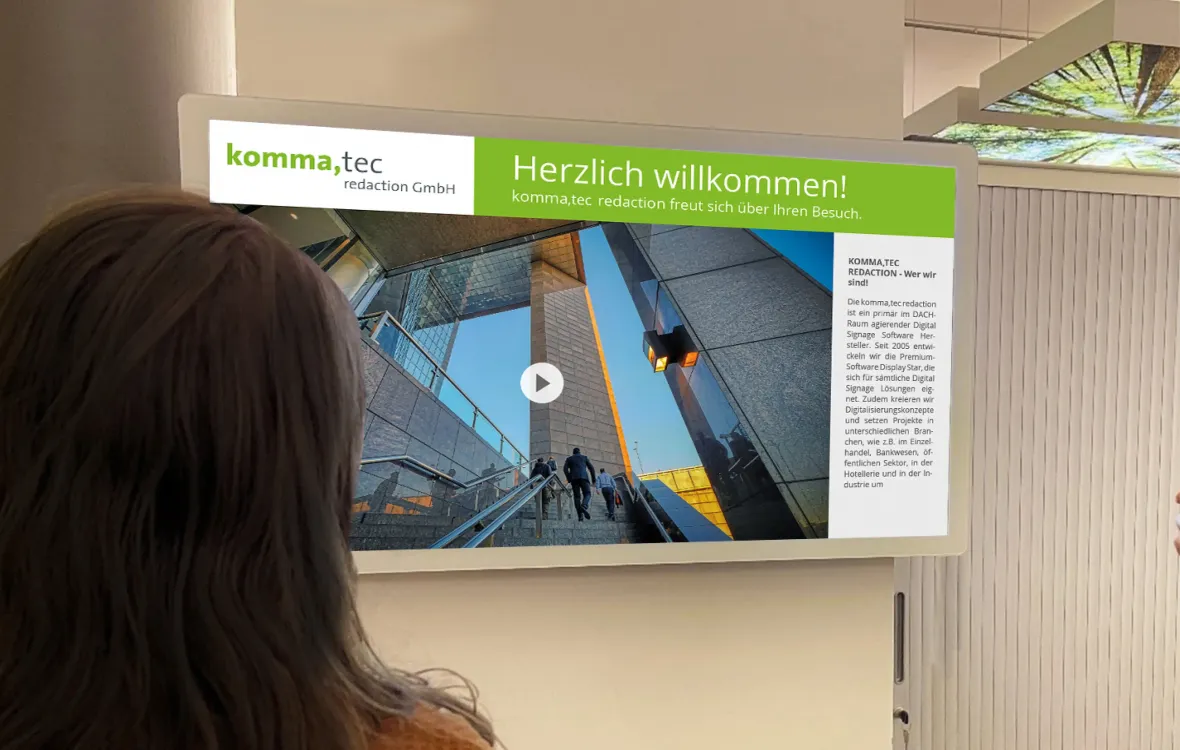 Eine Person betrachtet ein digitales Begrüßungsdisplay, das eine Nachricht der komma,tec redaction GmbH und ein Bild eines modernen Gebäudes zeigt.
