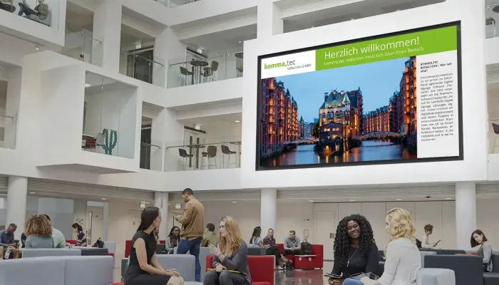 Ein großes digitales Infoboard zeigt Informationen in einem modernen, offenen Büro- und Aufenthaltsbereich, in dem Menschen auf Sitzgelegenheiten entspannen.