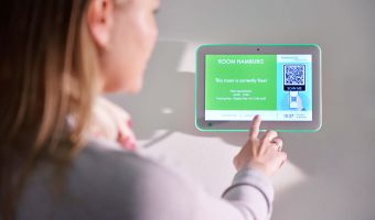 Eine Person interagiert mit einem digitalen Türschild mit Touch-Funktion, das aktuelle Raumverfügbarkeiten und einen QR-Code anzeigt.
