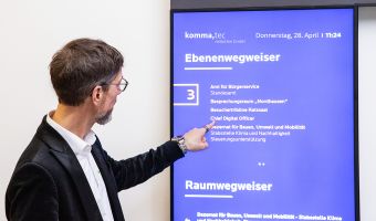 Ein Mann zeigt auf einen digitalen Ebenenwegweiser mit Informationen zu Räumen und Abteilungen, bereitgestellt von komma,tec redaction