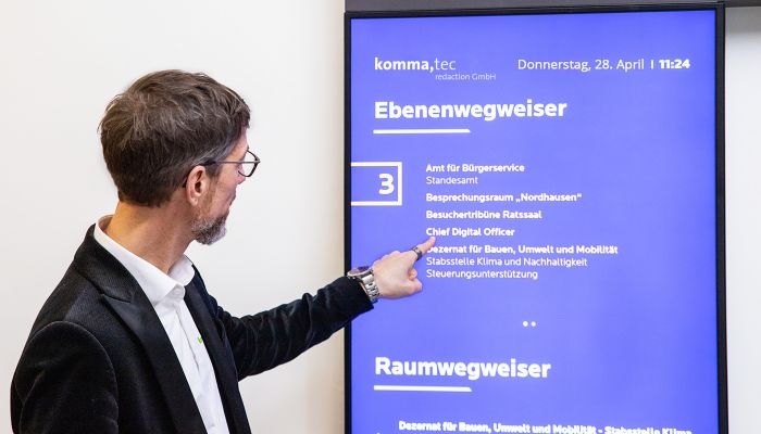 Ein Mann zeigt auf einen digitalen Ebenenwegweiser mit Informationen zu Räumen und Abteilungen, bereitgestellt von komma,tec redaction