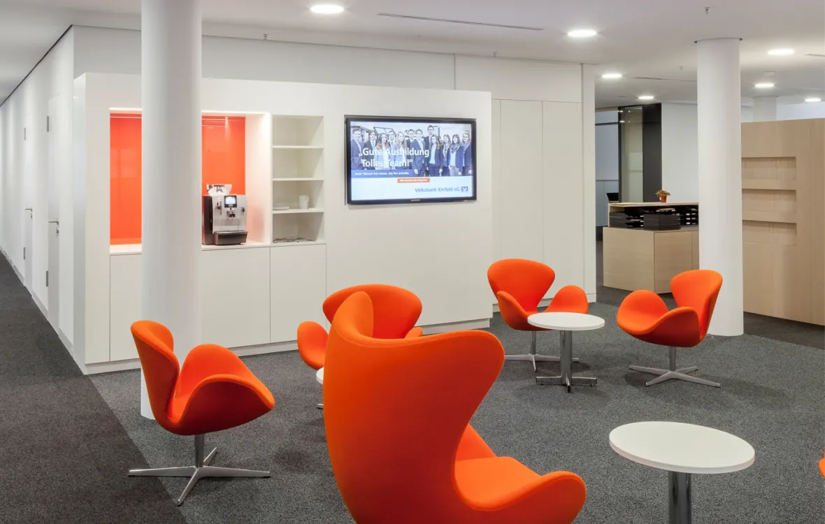 Ein stilvoller Wartebereich mit orangefarbenen Sesseln und einem digitalen Display, das Informationen über eine Bank anzeigt.