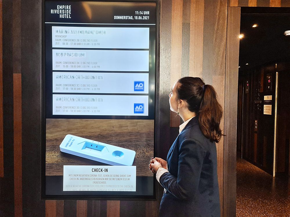 Frau betrachtet einen digitalen Begrüßungsbildschirm im Empire Riverside Hotel, der Veranstaltungsinformationen und Check-in-Hinweise anzeigt.