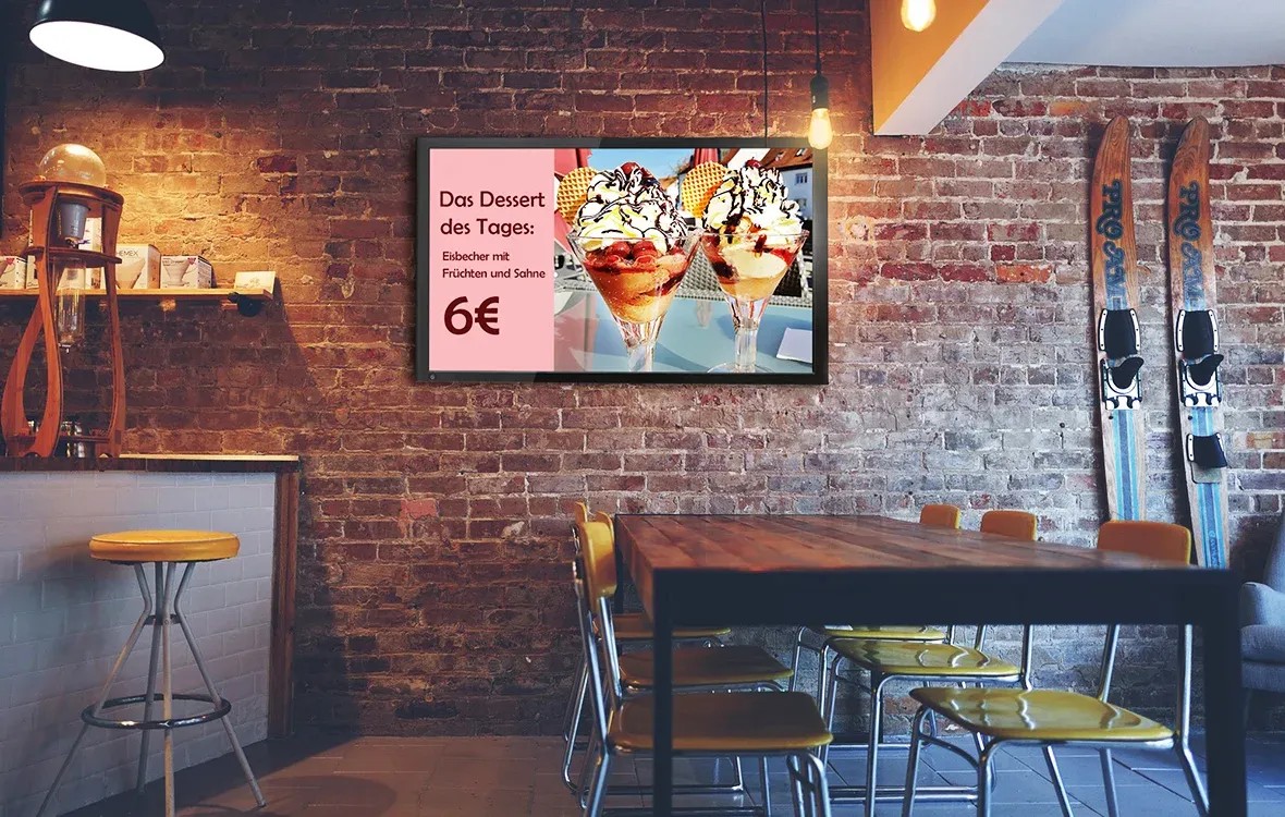 Ein gemütliches Café mit einer Ziegelwand, an der ein digitaler Bildschirm hängt. Der Bildschirm zeigt das Angebot 'Das Dessert des Tages: Eisbecher mit Früchten und Sahne, 6€' mit einem Bild von zwei Eisbechern.