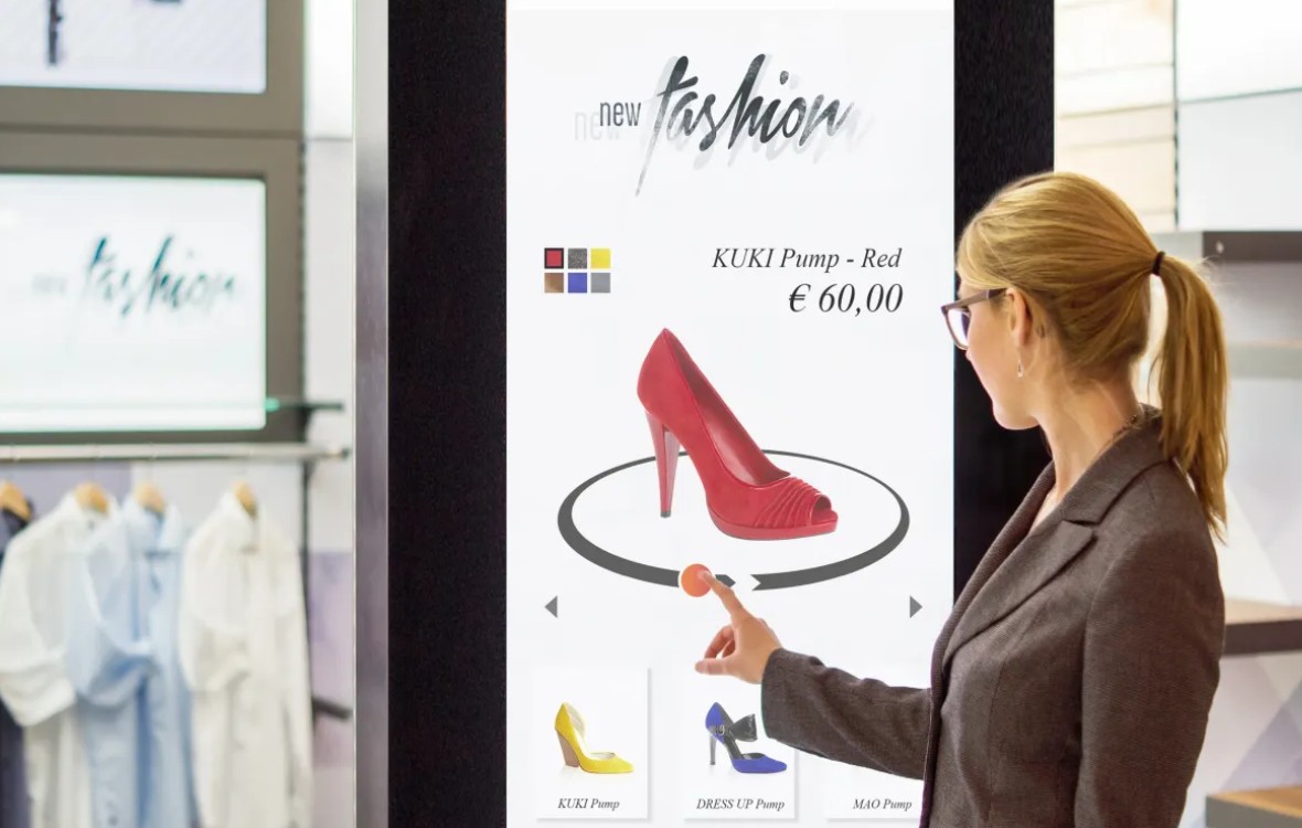 Frau interagiert mit einem interaktiven Touch-Display am Point of Sale, das ein rotes KUKI Pump-Modell fĂŒr 60 ⏠anzeigt, mit Auswahlmöglichkeiten fĂŒr andere Farben und Modelle.